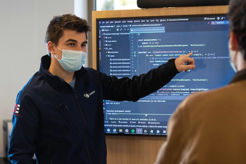 A person wearing a mask points to a screen with code on it